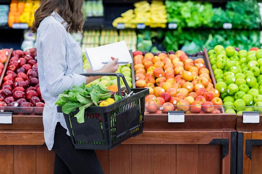 A woman is shopping in a grocery store in the produce department. In won hand, she carries a black basket on her arm and and pen and notepad in her hand.