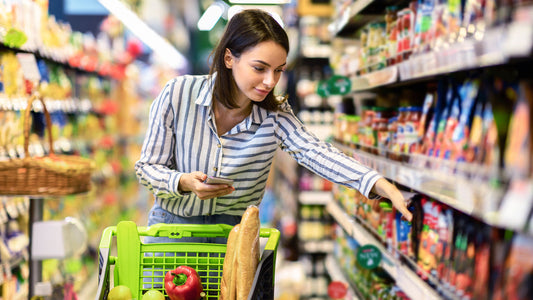 A women pushing a shopping cart in a grocery store and grabbing an item from the shelf beside her. 
